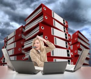 Stressed young woman using multiple computers, with piles of folders on the backgrounds